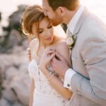groom-kisses-bride-hugging-her-shoulders-on-the-rocky-seashore.jpg