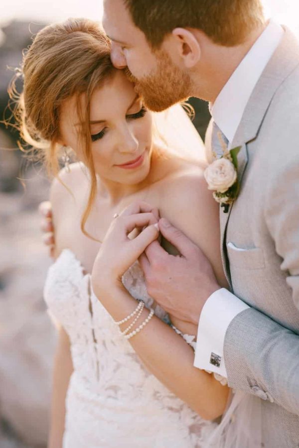 groom-kisses-bride-hugging-her-shoulders-on-the-rocky-seashore.jpg